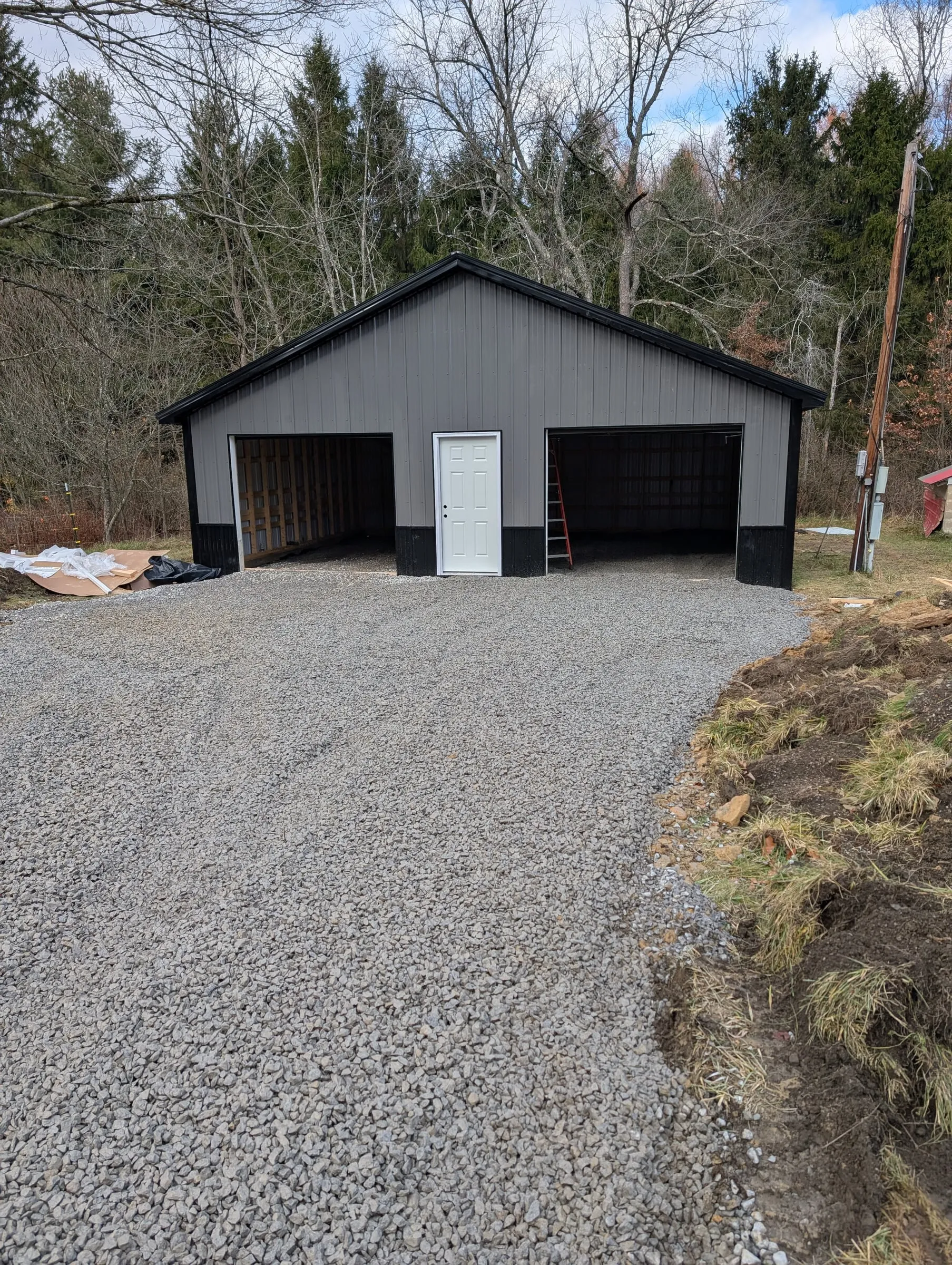 Garage sided in gray and black steel with door openings before the overhead doors went in, gravel drive in front